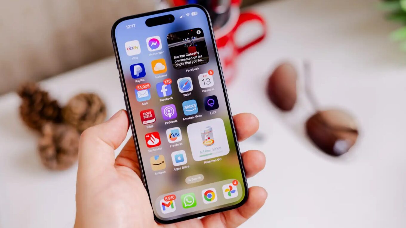 HyperOS 2.0 x iOS 26: Qual Sistema Leva a Melhor em Animações e Personalização? 1 Mão segurando um iPhone com tela inicial exibindo diversos ícones de aplicativos e um widget de foto, representando a interface do iOS 26 beta.