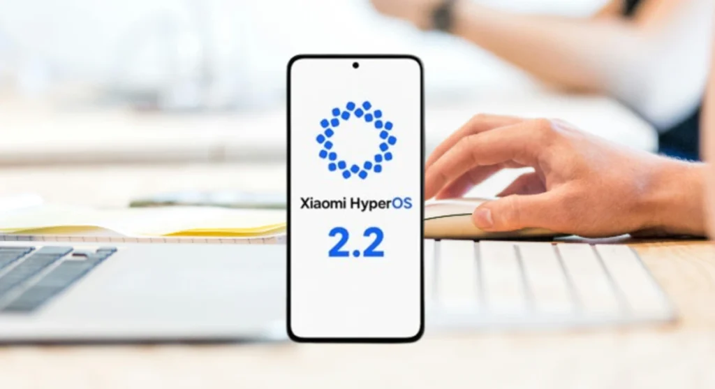 Celular mostrando o logo do Xiaomi HyperOS 2.2 sobre uma mesa de trabalho com teclado, mouse e laptop parcialmente visíveis, com uma pessoa usando o mouse ao fundo.
