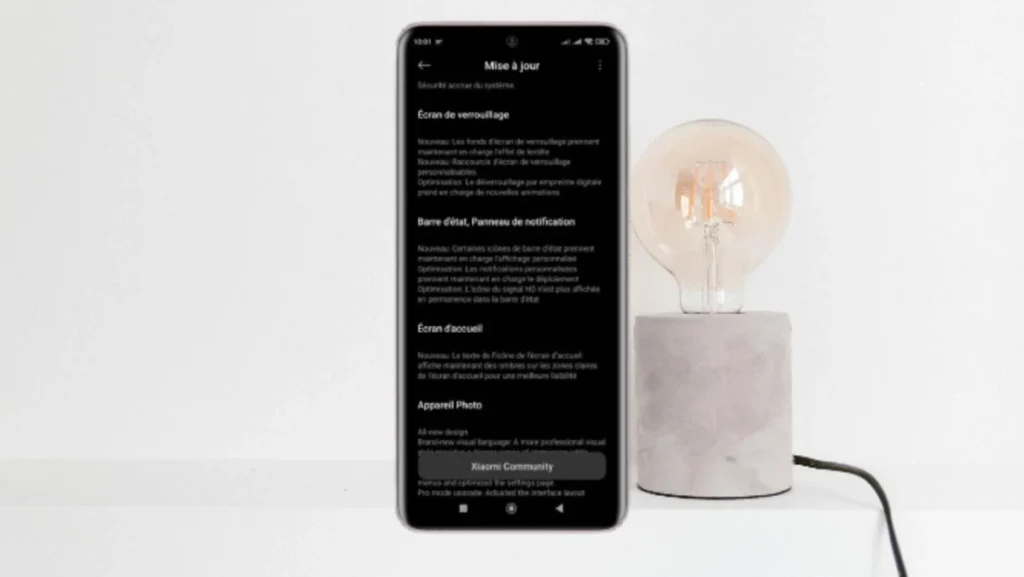 Smartphone mostrando a tela de atualização do HyperOS 2.2 com detalhes em francês, ao lado de uma luminária.