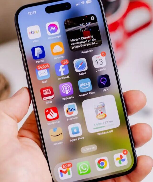 Mão segurando um iPhone com tela inicial exibindo diversos ícones de aplicativos e um widget de foto, representando a interface do iOS 26 beta.