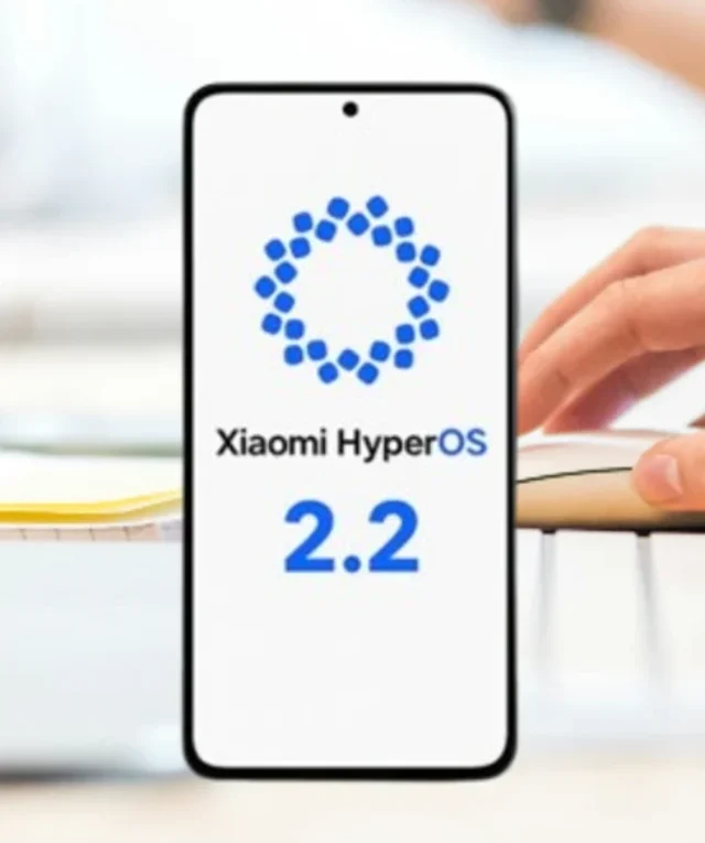 Celular mostrando o logo do Xiaomi HyperOS 2.2 sobre uma mesa de trabalho com teclado, mouse e laptop parcialmente visíveis, com uma pessoa usando o mouse ao fundo.