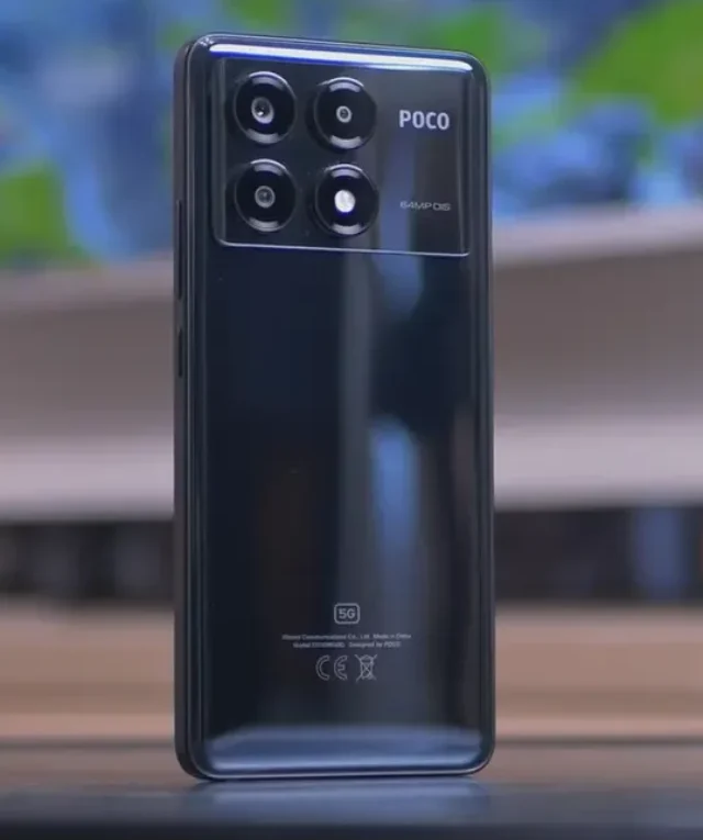 Smartphone POCO X6 Pro na cor preta, exibindo a parte traseira com o módulo da câmera retangular, que possui quatro lentes circulares e as inscrições 'POCO' e '64MP OIS', sobre uma superfície de madeira e fundo desfocado.