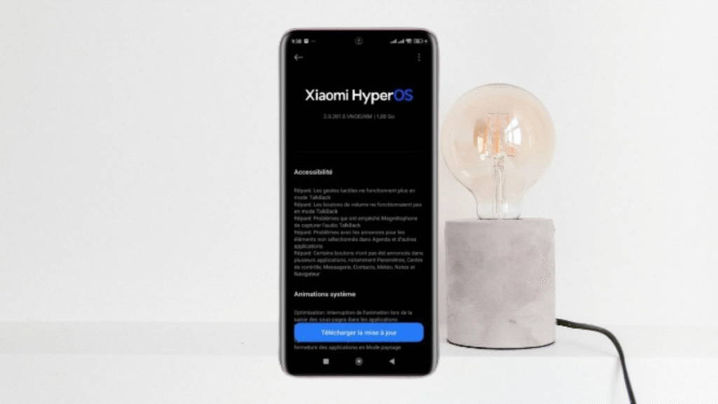 Smartphone Xiaomi com a tela exibindo as notas de atualização do HyperOS 2.0.201, focado em melhorias de acessibilidade e animações, ao lado de uma lâmpada de mesa.