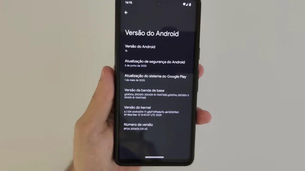 Uma mão segura um smartphone que exibe a tela "Versão do Android". A tela mostra "Versão do Android 16", "Atualização de segurança do Android 5 de junho de 2025" e "Atualização do sistema do Google Play 1 de maio de 2025", além de informações sobre a versão da banda de base, versão do kernel e número da versão.