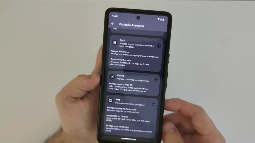 Duas mãos seguram um smartphone exibindo a tela "Proteção Avançada". Várias opções de segurança são listadas, incluindo "Apps" com proteção contra bugs e apps perigosos, "Google Play Protect" para verificar apps e malware, "Apps desconhecidos" para bloquear instalação de fontes desconhecidas, "Redes" para impedir conexões sem segurança, "Web" para proteger contra sites perigosos e opções relacionadas à navegação e proteção JavaScript.