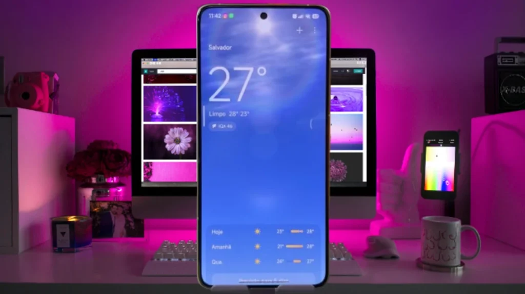 Smartphone Xiaomi exibindo o aplicativo do Clima do HyperOS 3 com a temperatura de 27 graus em Salvador, Brasil, e previsão para os próximos dias, em um ambiente com iluminação roxa.