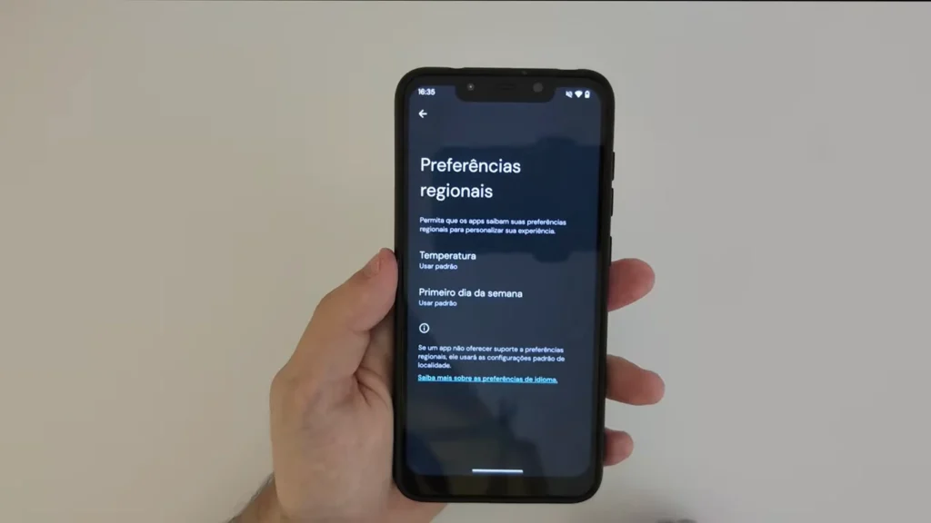 Uma mão segura um smartphone que exibe a tela de configurações "Preferências regionais". As opções visíveis incluem "Temperatura" e "Primeiro dia da semana", ambas configuradas como "Usar padrão". Há uma descrição explicando que os apps podem usar essas preferências para personalizar a experiência e uma nota sobre o comportamento de apps que não oferecem suporte a preferências regionais.