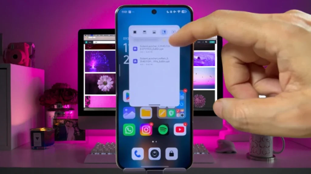 Texto Alt: Um celular Android moderno exibe uma janela sobreposta mostrando dois arquivos de instalação de launcher (HyperOS Launcher 2). Um dedo aponta para o botão de instalação do arquivo selecionado. A tela inicial do celular com diversos aplicativos coloridos é visível abaixo da janela. Ao fundo, uma mesa de escritório com um computador iMac exibindo papéis de parede abstratos e florais em sua tela, um teclado branco, um mouse branco, uma xícara branca e outros objetos decorativos como uma câmera rosa e um vaso de flores roxas são visíveis em um ambiente com iluminação rosa e roxa.