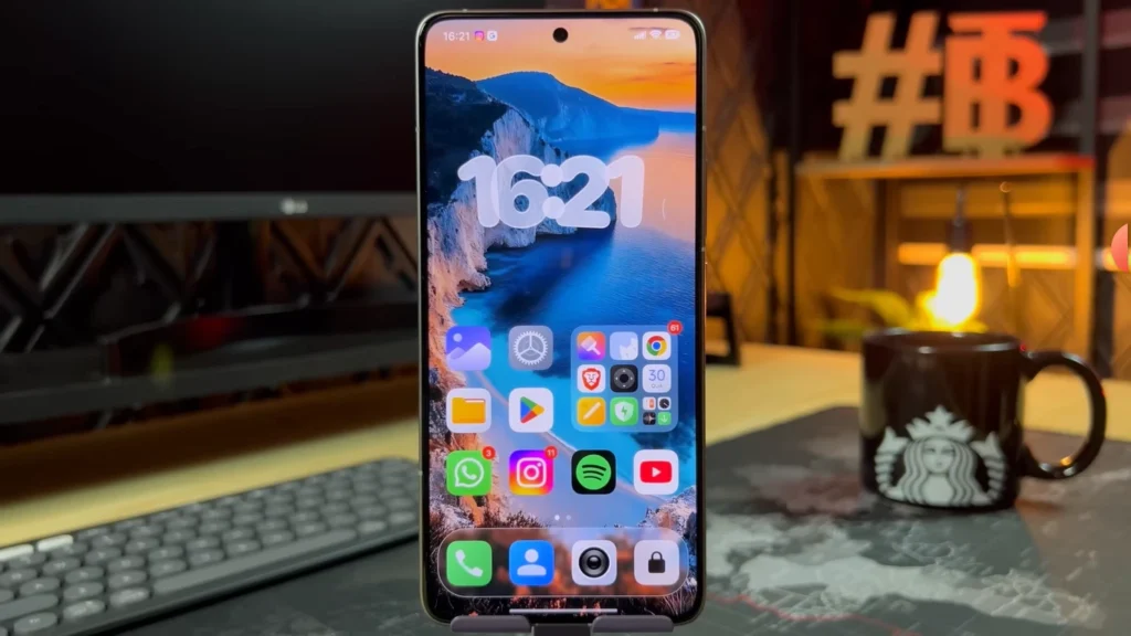 Smartphone Xiaomi exibindo a tela inicial com o horário 16:21, ícones de aplicativos organizados e um papel de parede de paisagem de praia e montanhas, em um ambiente de escritório.