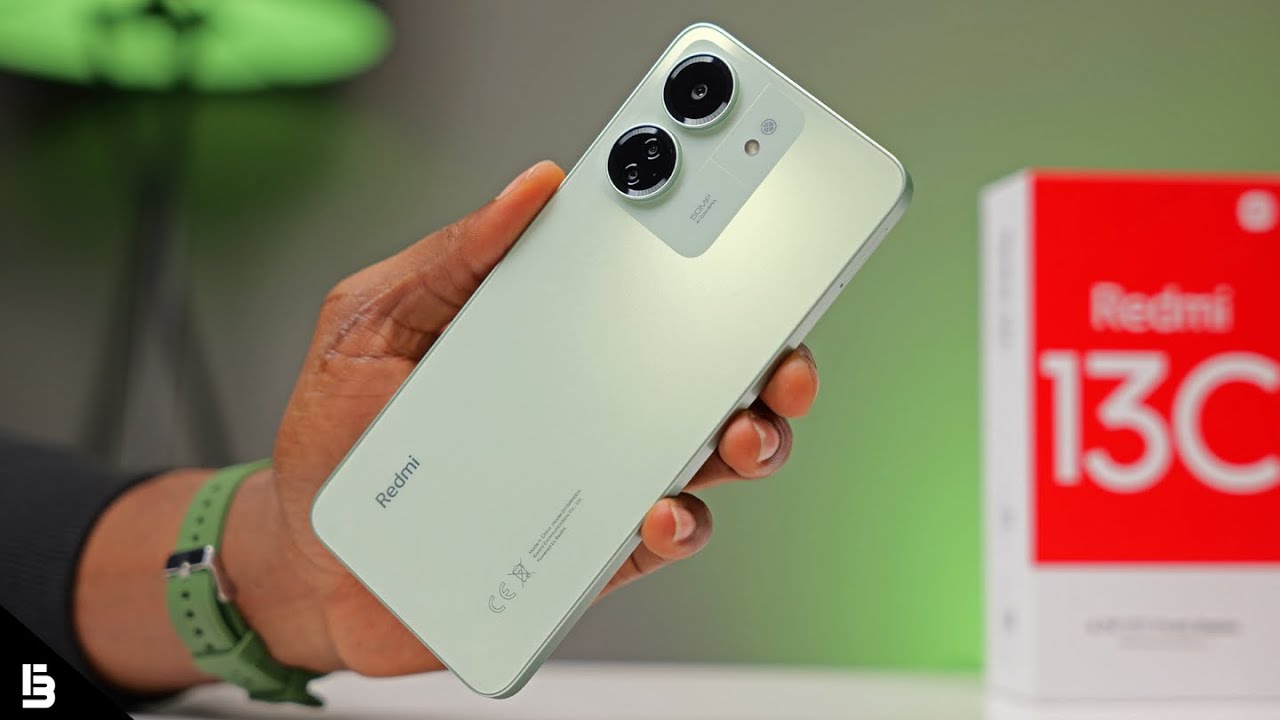 Um close-up de um smartphone Redmi 13C na cor verde-claro sendo segurado na mão de uma pessoa, com o logo da Redmi visível na parte inferior e um módulo de câmera dupla na parte superior. Ao fundo, parcialmente visível, está a caixa vermelha do Redmi 13C.
