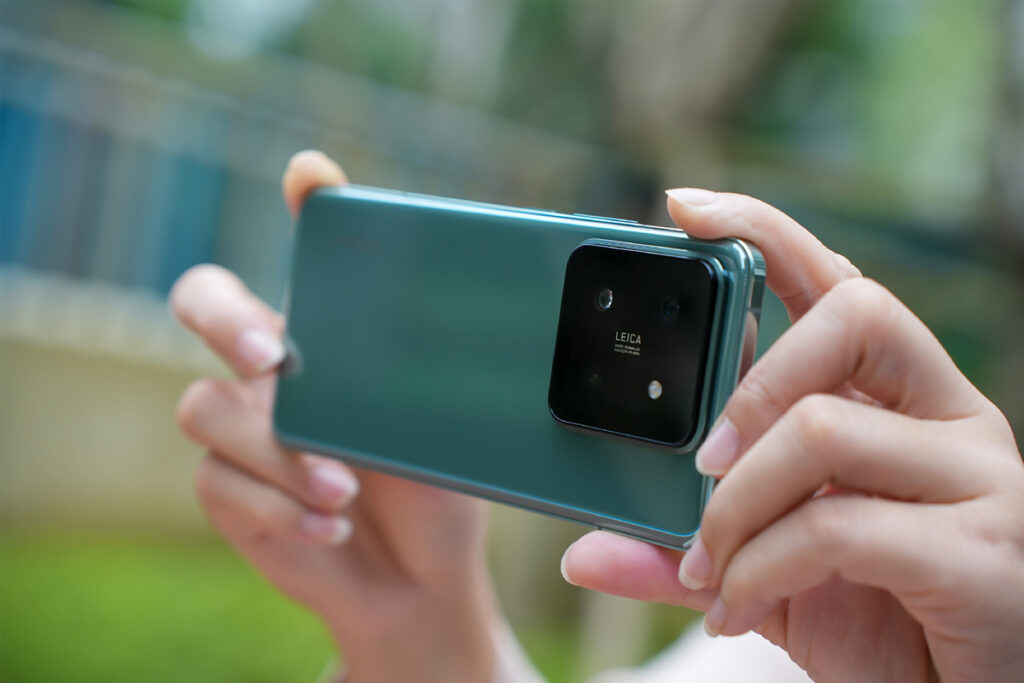 Mãos segurando horizontalmente a parte traseira de um smartphone Xiaomi14 Pro verde, com um módulo de câmera preto proeminente que inclui a marca Leica e múltiplos sensores.