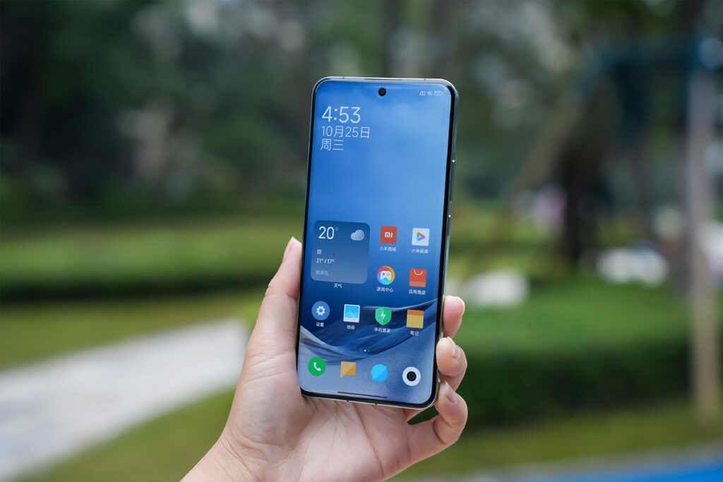 Mão segurando verticalmente um smartphone Xiaomi 14 Pro com a tela inicial do HyperOS visível, exibindo widgets de clima e data, e ícones de aplicativos em um fundo de tela de paisagem.