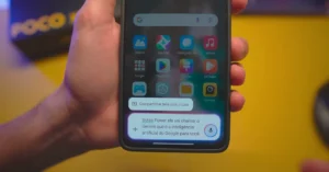 Mão segurando um smartphone Xiaomi com HyperOS 2.2 exibindo a tela inicial com um pop-up do Google Assistente, mencionando o Gemini como a inteligência artificial do Google.
