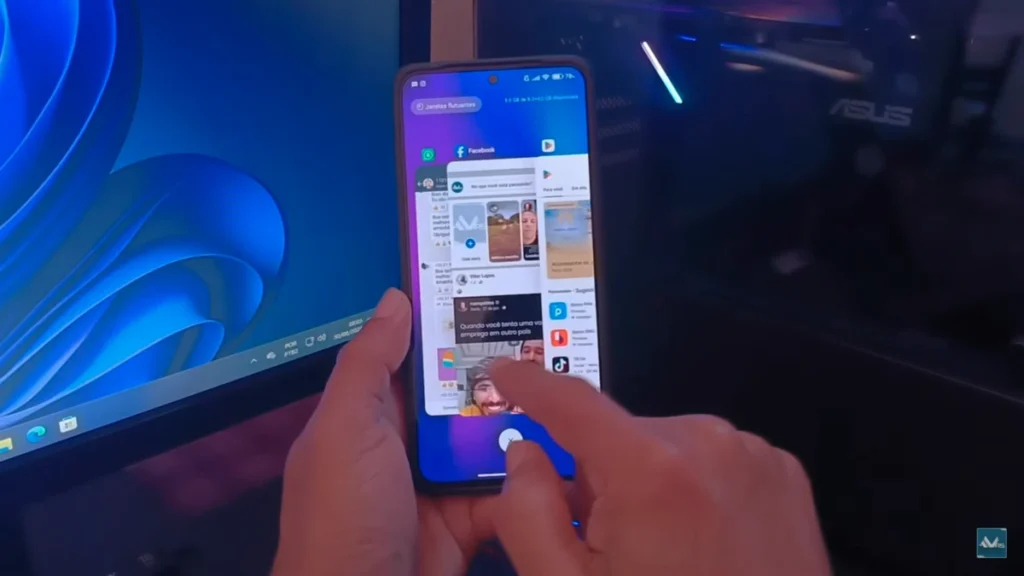 A imagem mostra em destaque um Redmi Note 13 4G com o novo menu de apps recentes do Xiaomi HyperOS 3, que adota um estilo iOS. Várias janelas de aplicativos, como Facebook e outras redes sociais, são visíveis na tela, organizadas em um formato de grade ou cascata, indicando a facilidade de navegação entre os apps abertos. Uma mão segura o aparelho, interagindo com o sistema. Ao fundo, um ambiente de escritório moderno com iluminação em tons de azul e verde complementa a cena, com uma mesa escura, um tripé para celular, um monitor e outros equipamentos eletrônicos, criando uma atmosfera tecnológica. A interface do Xiaomi HyperOS 3 promete otimização e uma experiência de usuário aprimorada, especialmente para quem busca a organização e a estética do sistema da Apple.
