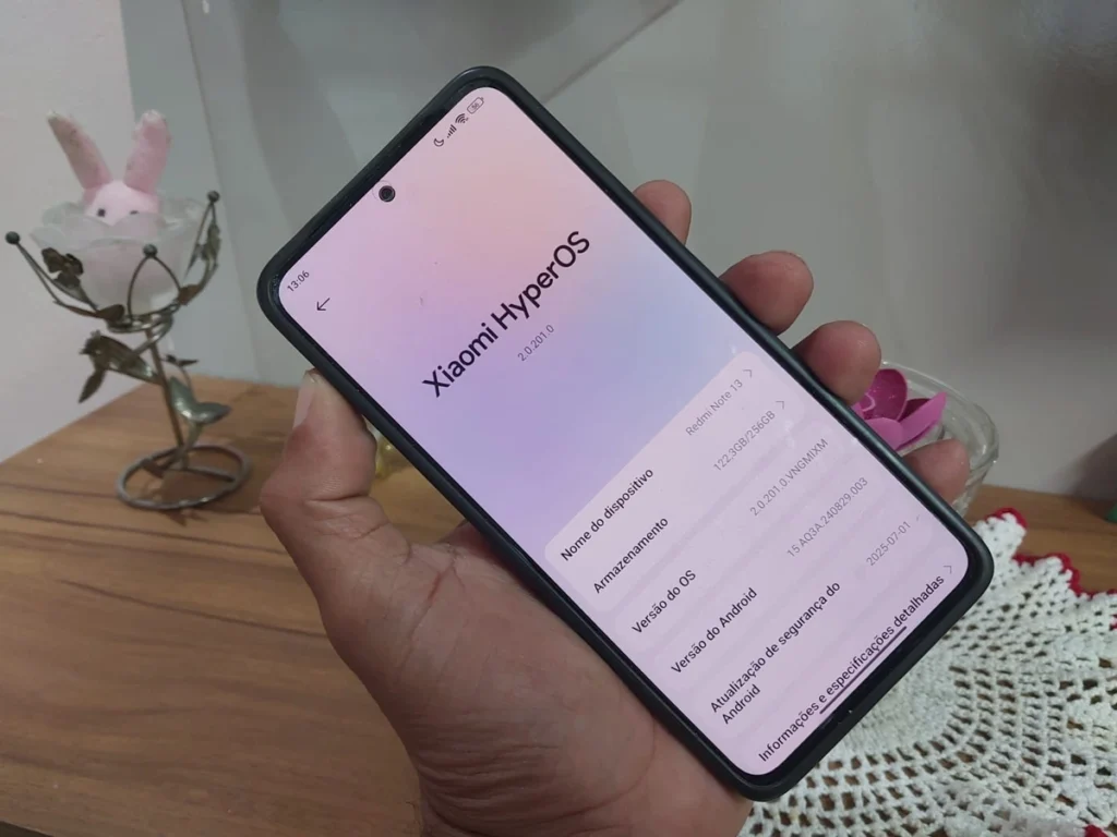 Mão segurando um smartphone Xiaomi Redmi Note 13 com a tela exibindo as informações do sistema **HyperOS 2.0.201.0**, incluindo a versão do Android e a atualização de segurança de julho de 2025.