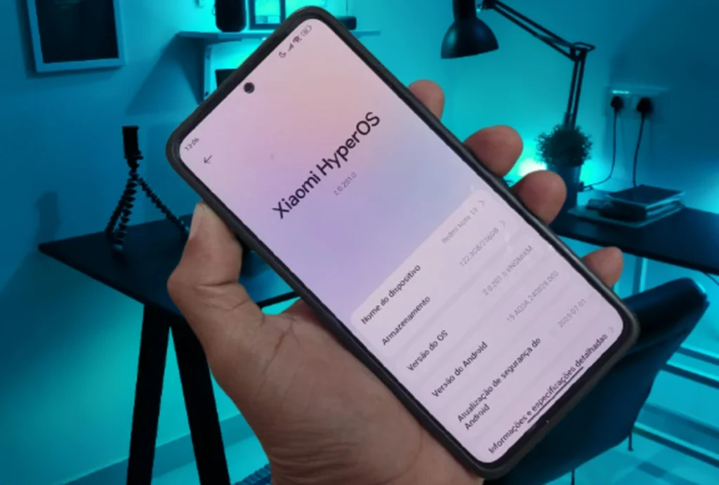 Mão segurando um smartphone Xiaomi Redmi Note 13 com a tela exibindo as informações do sistema **HyperOS 2.2**, incluindo a versão do Android e a atualização de segurança de julho de 2025.