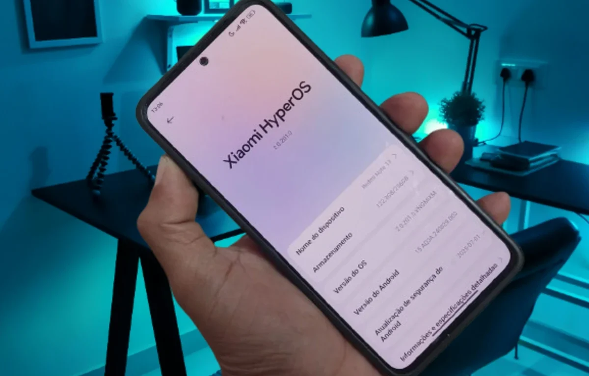 Mão segurando um smartphone Xiaomi Redmi Note 13 com a tela exibindo as informações do sistema **HyperOS 2.2**, incluindo a versão do Android e a atualização de segurança de julho de 2025.