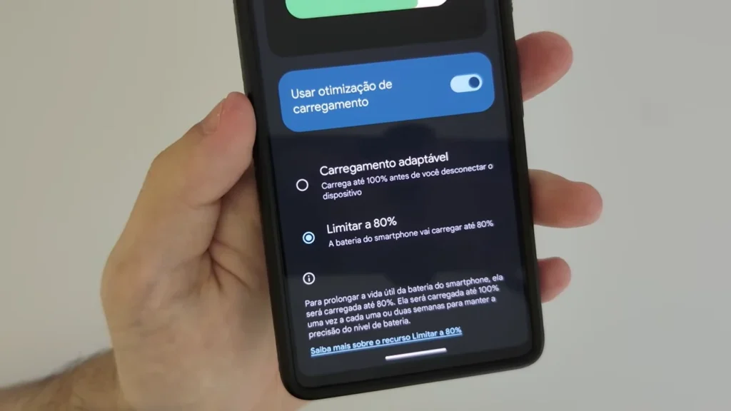 Uma mão segura um smartphone que exibe as configurações de bateria, com foco nas opções de carregamento. A tela mostra um toggle para "Usar otimização de carregamento" (ativado), e duas opções de carregamento adaptativo: "Carregamento adaptável" e "Limitar a 80%" (selecionada). Há também uma descrição sobre como limitar a 80% pode prolongar a vida útil da bateria, com um link para saber mais.