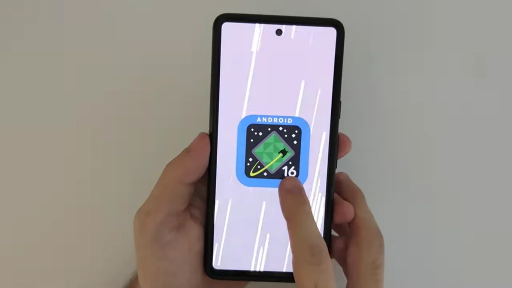Uma pessoa segura um smartphone preto na vertical, e a tela exibe o logo de inicialização do Android 16. O logo é um losango verde com um contorno amarelo dentro de um quadrado arredondado azul com bordas pretas e a palavra "ANDROID" no topo. O número "16" estilizado em amarelo está na parte inferior direita do losango. O fundo da tela é claro com linhas verticais em tom pastel.