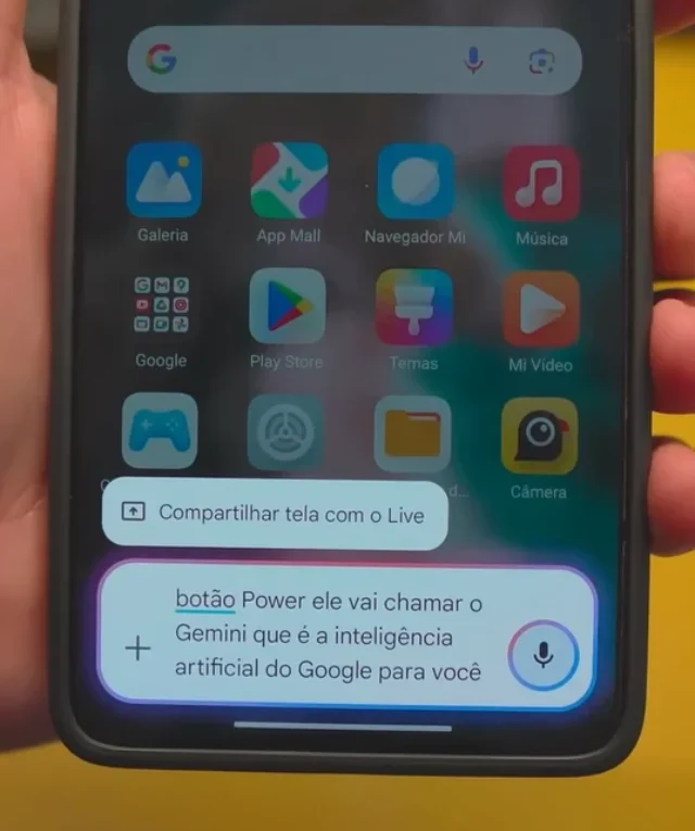 Mão segurando um smartphone Xiaomi com HyperOS 2.2 exibindo a tela inicial com um pop-up do Google Assistente, mencionando o Gemini como a inteligência artificial do Google.