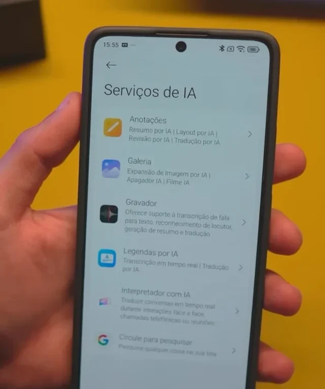 Mão segurando um smartphone Xiaomi exibindo a tela "Serviços de IA" do HyperOS 2.2, mostrando recursos como Anotações, Galeria, Gravador e Legendas.