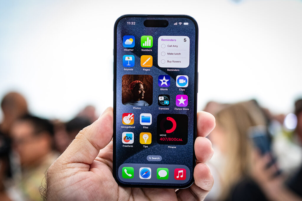 Mão segurando um iPhone 15 Pro Max em primeiro plano, com a tela inicial exibindo aplicativos e widgets coloridos, e um fundo desfocado de pessoas.
