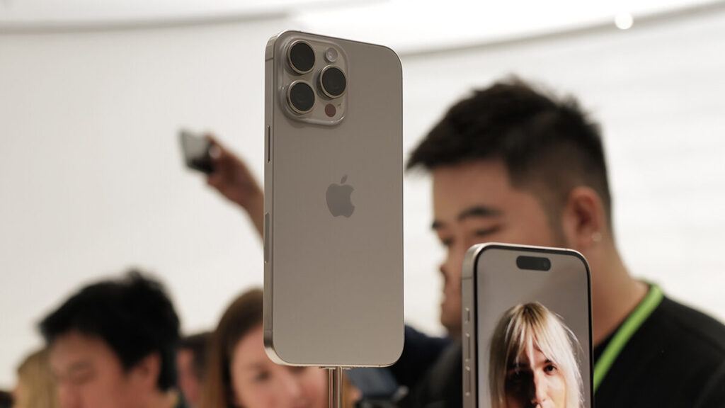 Parte traseira de um iPhone 15 Pro Max dourado em exibição, com seu sistema de câmera tripla em destaque, e pessoas borradas ao fundo observando.