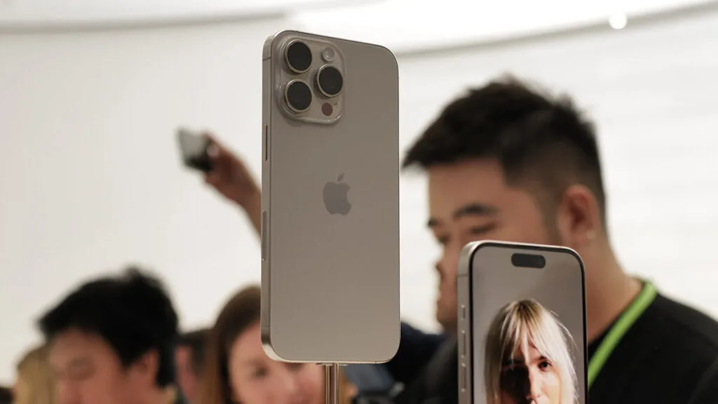 Parte traseira de um iPhone 15 Pro Max dourado em exibição, com seu sistema de câmera tripla em destaque, e pessoas borradas ao fundo observando.