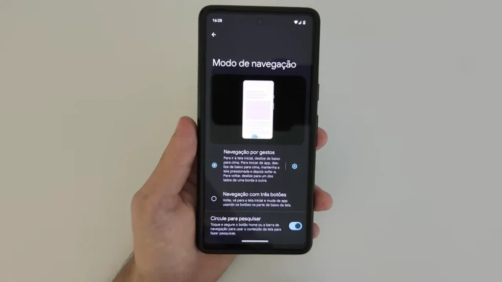 Uma mão segura um smartphone que exibe a tela de configurações "Modo de navegação". As opções visíveis são "Navegação por gestos" (selecionada), "Navegação com três botões" e "Círculo para pesquisar", com descrições detalhadas para cada método de navegação. Uma ilustração animada no topo da tela demonstra a navegação por gestos.