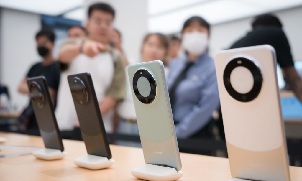 Uma fileira de smartphones com grandes módulos de câmera circulares, em cores preto, cinza e branco, expostos em suportes em uma loja ou evento. Pessoas borradas estão no fundo.
Smartphones Huawei em exibição, simbolizando os dispositivos que usam a tecnologia NearLink, adotada recentemente pela Xiaomi.