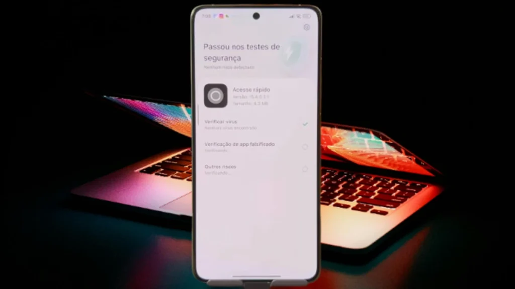 A imagem captura a tela de um smartphone mostrando o processo de verificação de segurança dos novos apps do HyperOS 2.2, indicando que passou nos testes. Um laptop com iluminação colorida aparece em segundo plano.