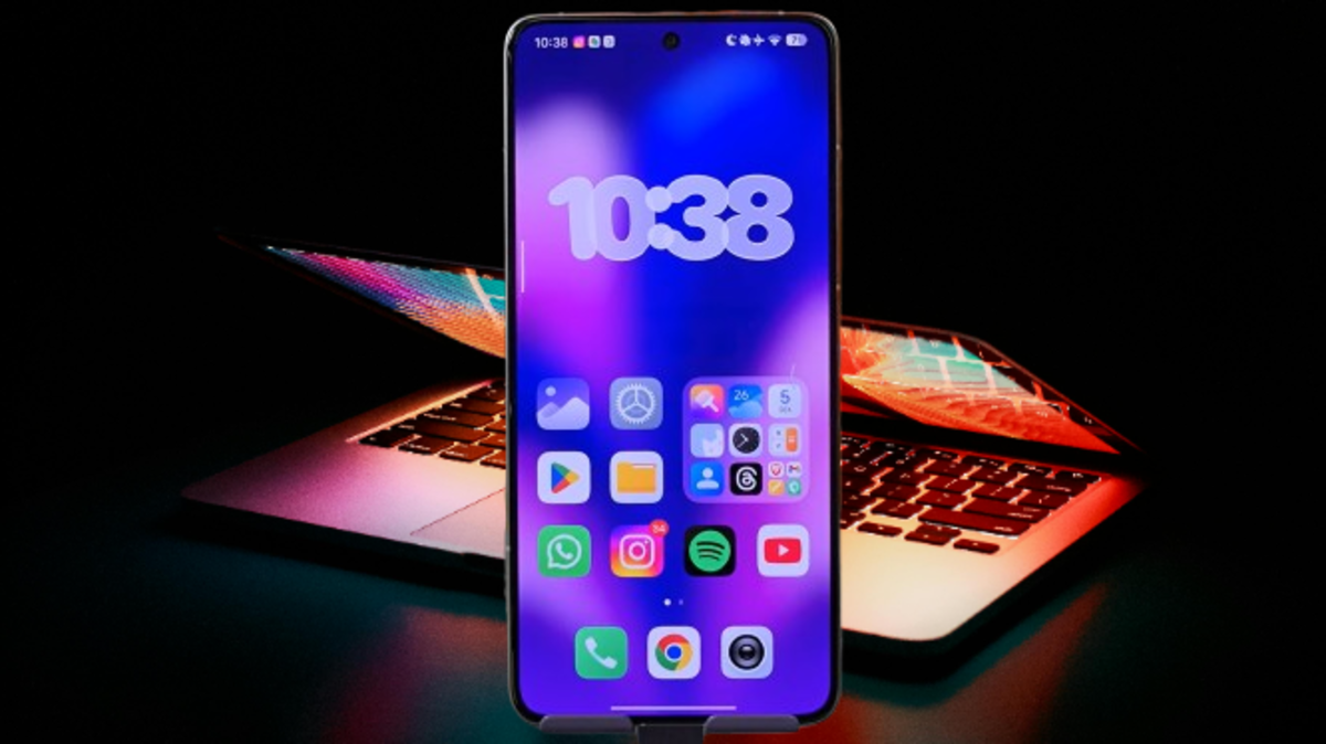 Um smartphone exibe a tela inicial do HyperOS Launcher 3, mostrando um papel de parede roxo-azulado, um widget de relógio grande e ícones de aplicativos, com um notebook com o teclado retroiluminado ao fundo.