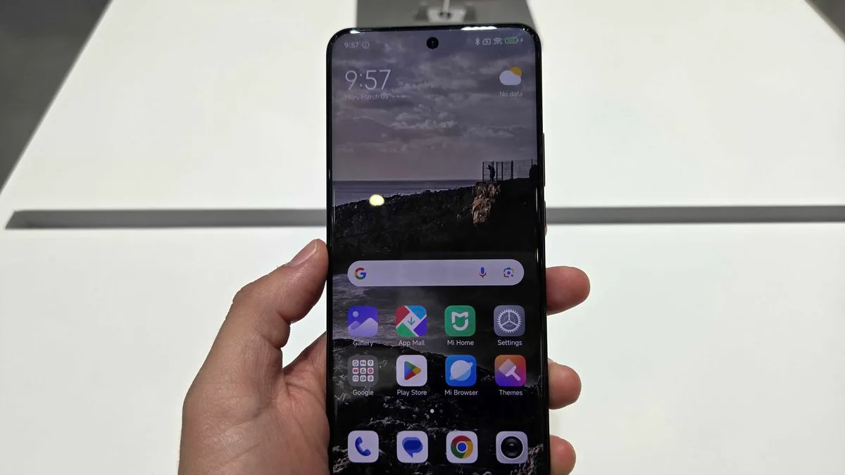 Mão segurando um smartphone Xiaomi exibindo a tela inicial com o sistema HyperOS 3. O papel de parede escuro (noturno) mostra uma paisagem de penhascos e nuvens, com ícones coloridos de aplicativos.