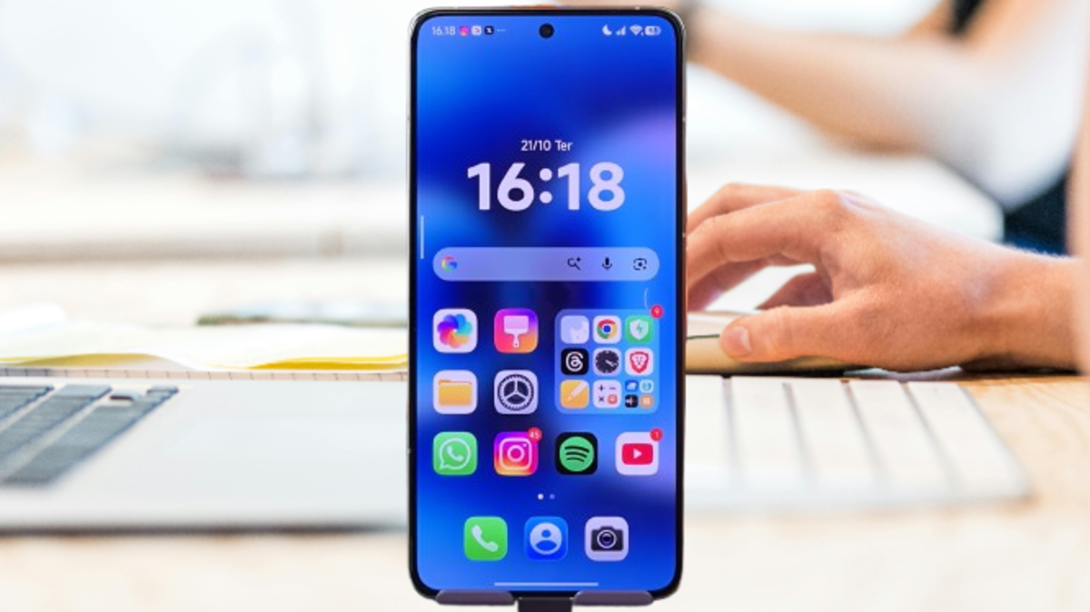 Smartphone exibindo a tela inicial com o HyperOS Launcher 3, apresentando widgets de hora e data e vários ícones de aplicativos coloridos, com uma pessoa trabalhando em um computador ao fundo.