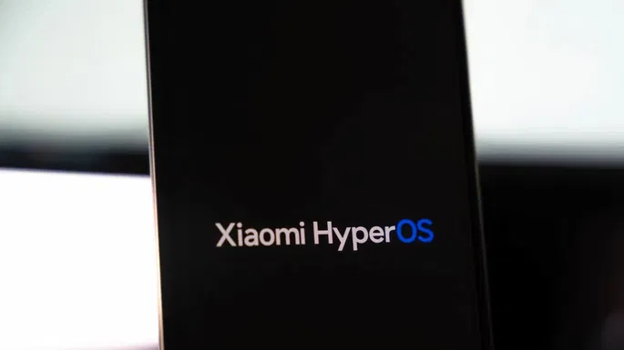 Esta imagem em close-up mostra um smartphone com a tela preta e o logo "Xiaomi HyperOS" centralizado. A palavra "Xiaomi" aparece em branco e "HyperOS" em um tom vibrante de azul, destacando a nova identidade visual do sistema. O fundo desfocado e claro sugere um ambiente interno, talvez um laboratório ou estúdio, realçando o foco no display do aparelho. A imagem evoca a expectativa pelo Xiaomi HyperOS 3, a nova aposta da Xiaomi em seu ecossistema.