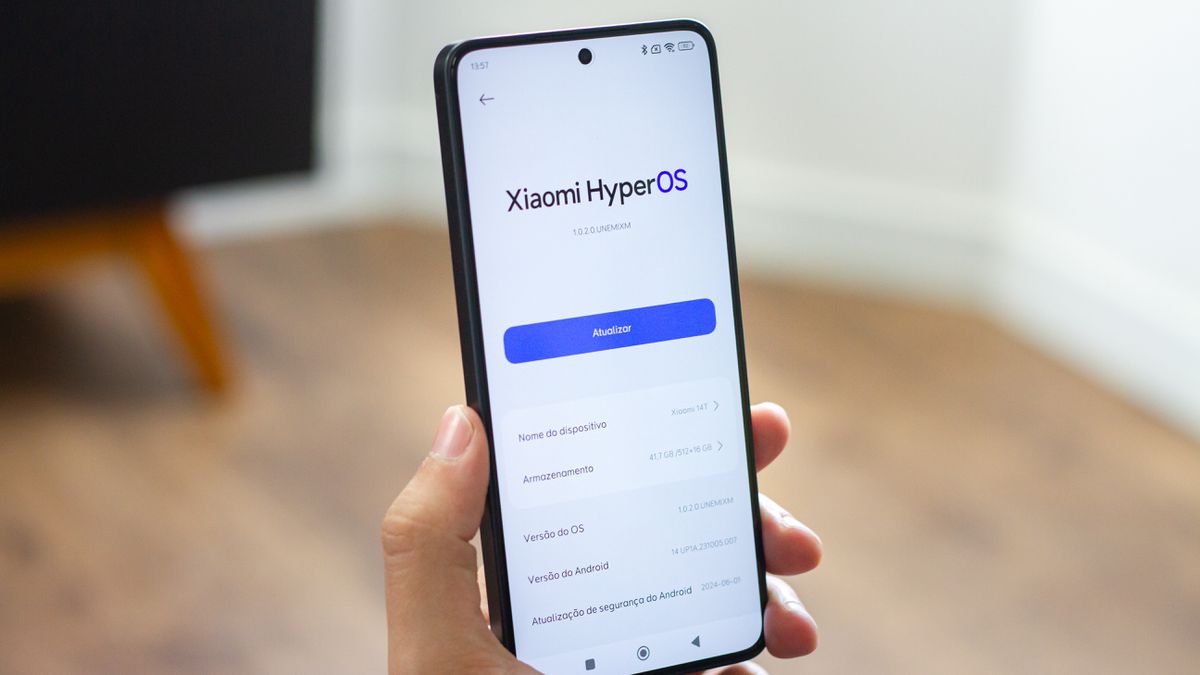 Uma mão segurando um smartphone POCO F6 Pro exibindo a tela de configurações do sistema. Na tela, é visível o logotipo do Xiaomi HyperOS e o botão azul 